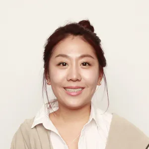 머리를 묶고 흰색 셔츠와 베이지색 가디건을 입은 채 미소 짓고 있는 여성