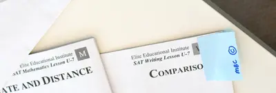 Two Elite Educational Institute workbooks on a desk: "SAT Mathematics Lesson U-7, Rate and Distance" and "SAT Writing Lesson U-7, Comparison," with a blue sticky note.