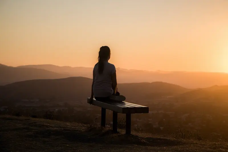 A person sits alone on a bench on a hilltop, watching the sun set over distant mountains