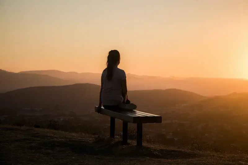 A person sits alone on a bench on a hilltop, watching the sun set over distant mountains