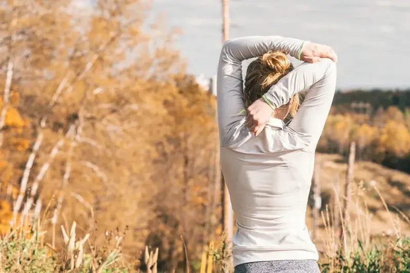 A woman seen from behind stretching her arms overhead outdoors among autumn trees
