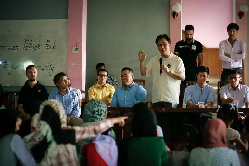 A man speaks into a microphone to a seated audience in a hall, with others seated behind him near a wall reading "Patrick Park".