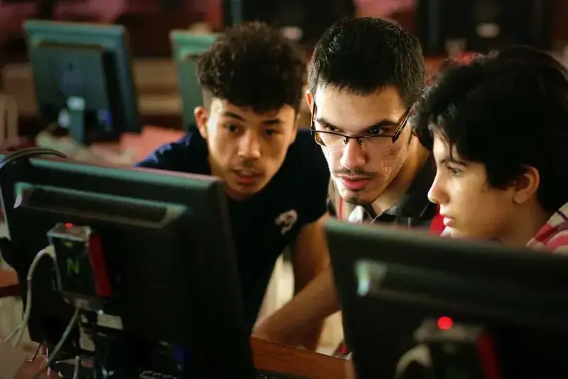 Three young students lean in together, looking intently at a computer monitor in a classroom lab.