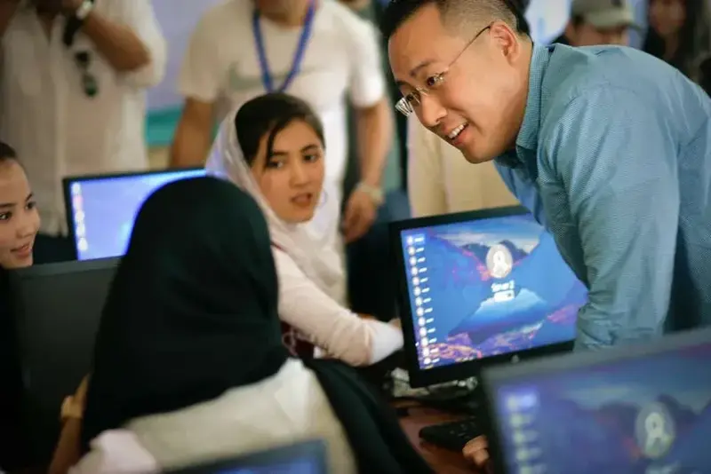A smiling man in a blue shirt leans over to talk with students working at desktop computers in a classroom.