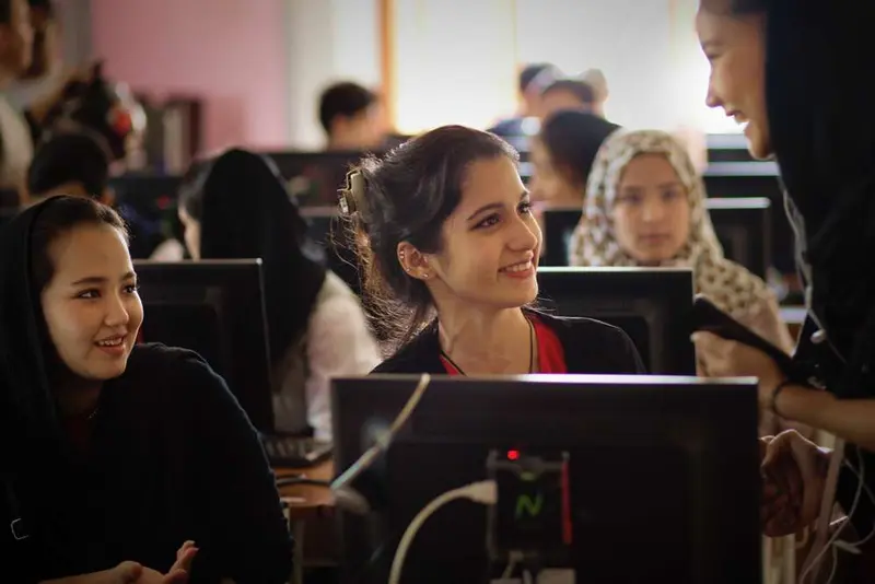 Smiling young women at computer workstations in a classroom, talking with a standing instructor.
