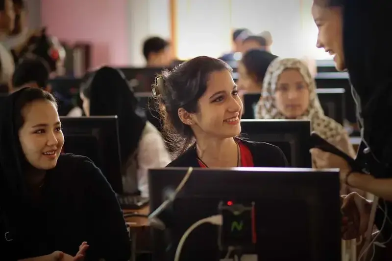 Smiling young women at computer workstations in a classroom, talking with a standing instructor.