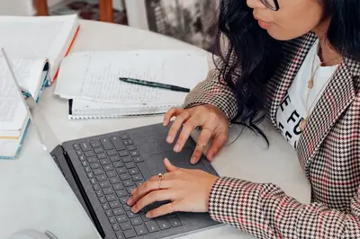 Student in a houndstooth blazer typing on a laptop beside stacks of notes, papers, and a pen