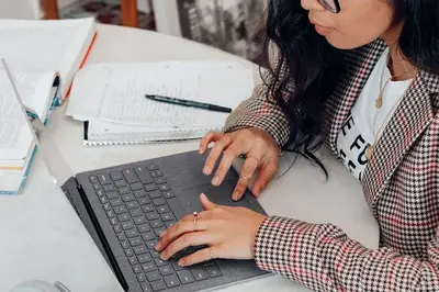 Student in a houndstooth blazer typing on a laptop beside stacks of notes, papers, and a pen
