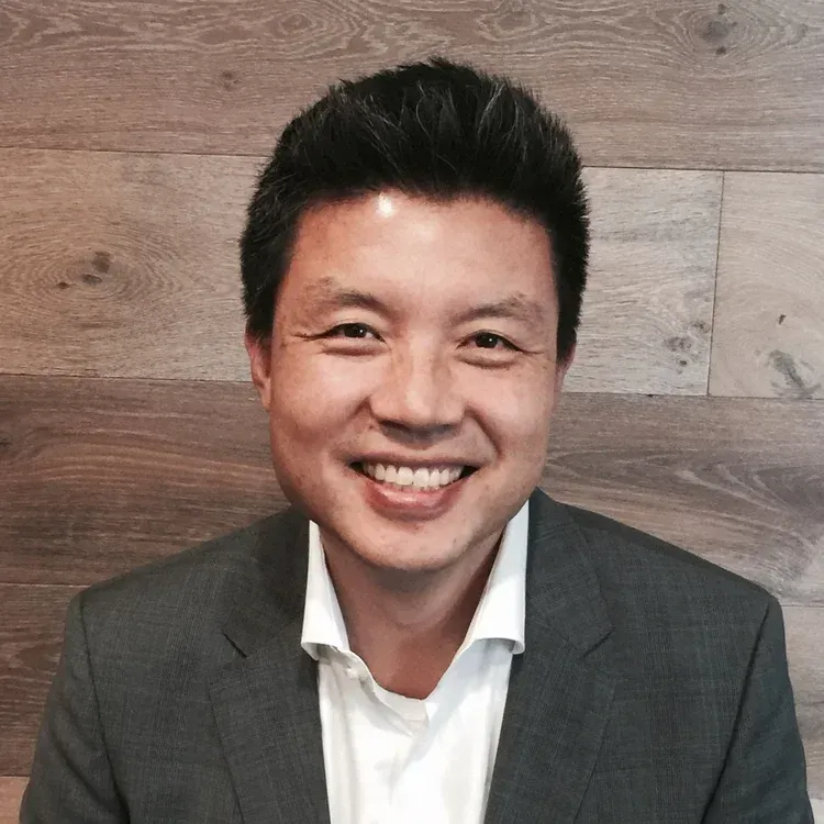 Smiling man in a gray blazer and white shirt in front of a wood plank wall