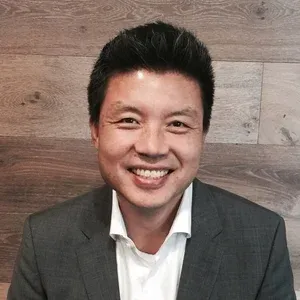 회색 재킷과 흰 셔츠를 입고 나무 벽 앞에서 미소 짓고 있는 남성
