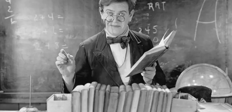 Vintage black-and-white photo of a smiling teacher in round glasses and bow tie holding an open book behind a row of books, with arithmetic problems on the chalkboard behind him