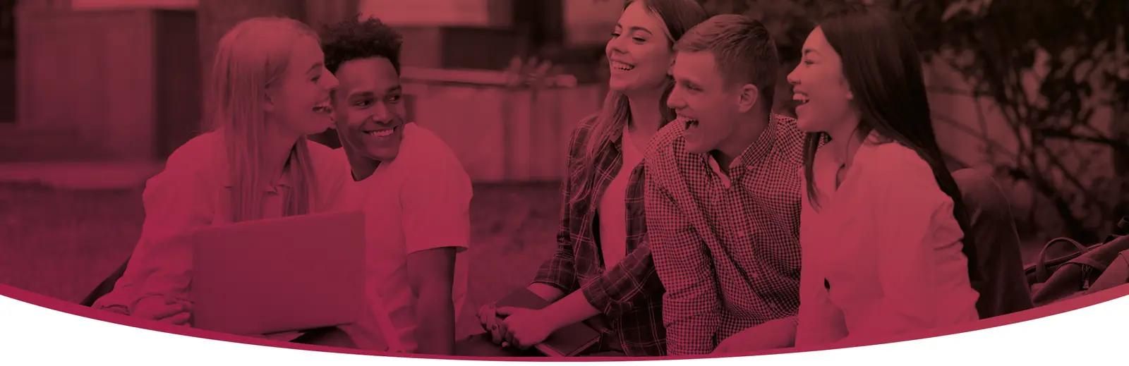 A group of students sitting outdoors together, smiling and talking around an open laptop