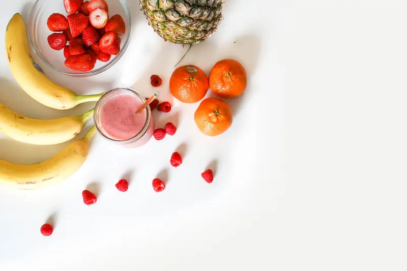Bananas, a bowl of strawberries, a pineapple, tangerines, raspberries and a pink smoothie on a white surface