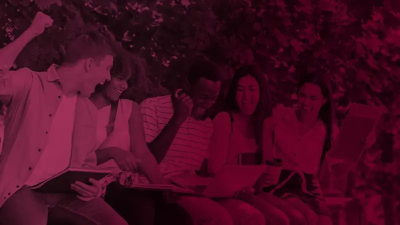 A group of smiling teenagers sitting outdoors together, looking at a laptop and notebooks.