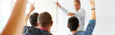 Students raise their hands in a classroom as a teacher at a whiteboard points to one of them