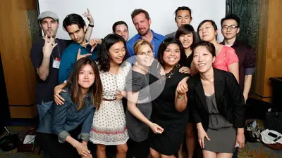 A group of smiling young students posing together, with a video play button overlaid on the photo.