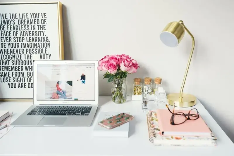 White desk with open laptop, pink roses, gold lamp, glasses on notebooks, and a framed motivational quote poster.