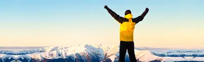 A hiker in a yellow jacket raises both arms in triumph atop a snow-covered mountain ridge at sunrise.