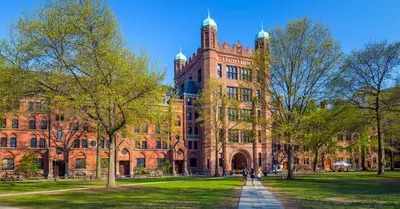 Students walking a path across a sunny green lawn toward a Gothic red-brick university building with green-domed towers.