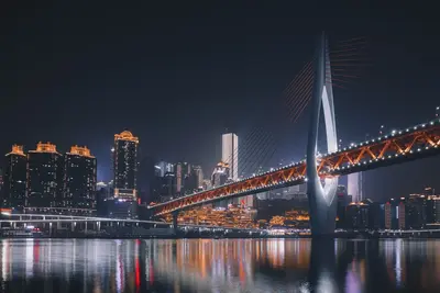 A city skyline at night, with a lit cable-stayed bridge reflected in the river below.