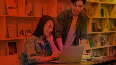 Two students smiling over a laptop in front of shelves of books