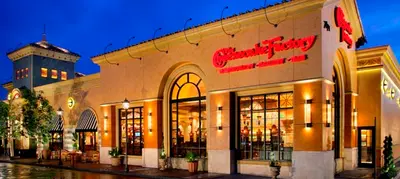 A Cheesecake Factory restaurant exterior lit up at dusk, with awnings, arched windows and palm trees out front.