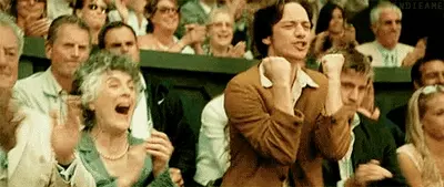 A crowd in stadium seats cheering, with a young man and an older woman clapping excitedly.