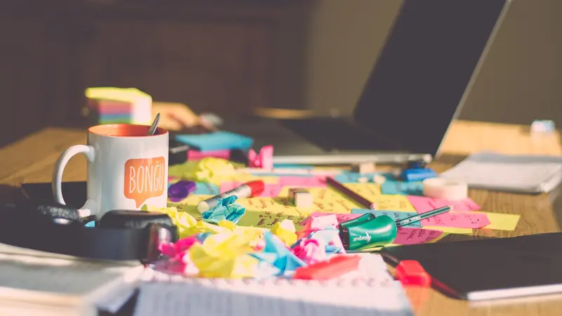 A cluttered desk covered in colorful sticky notes, markers, a notebook, a laptop, and a mug reading "Bongu".