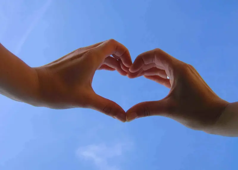 Two hands forming a heart shape against a clear blue sky