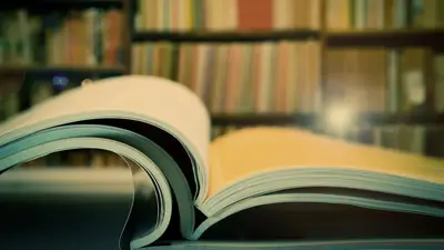 An open book on a table in a library, shelves of books behind it