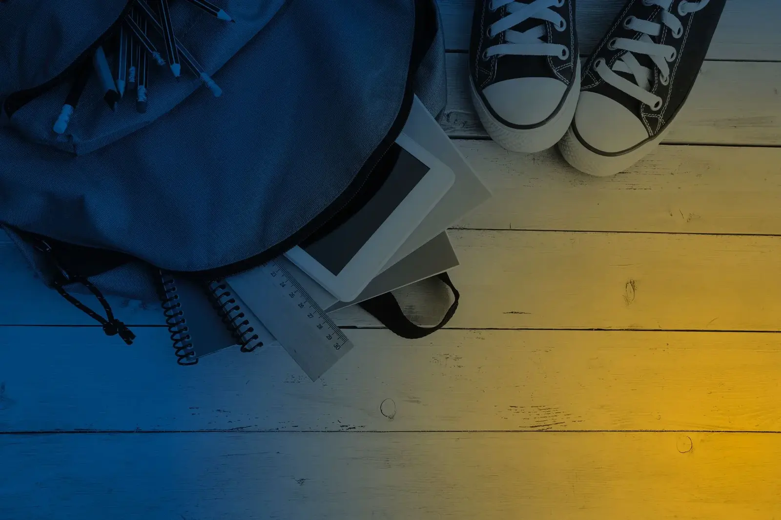 An open backpack with notebooks, pencils and a tablet beside a pair of sneakers on a wooden floor.