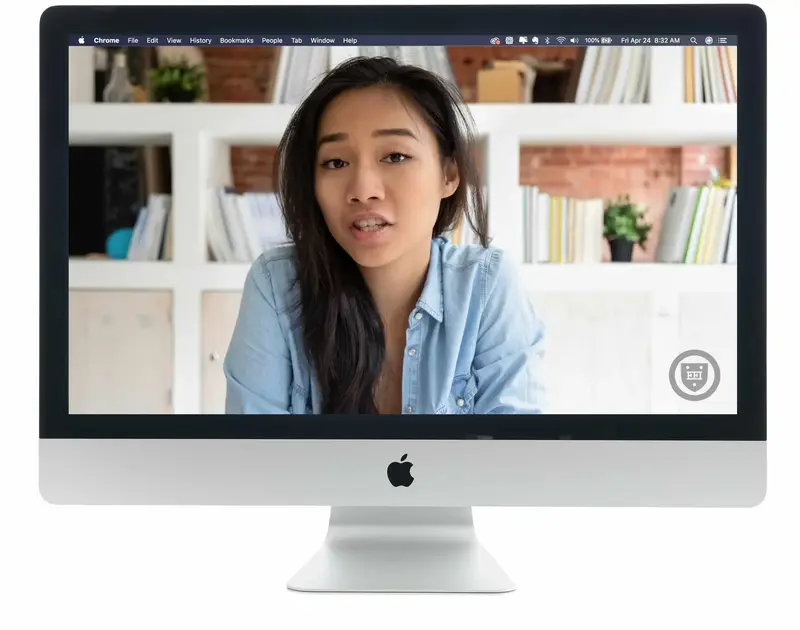 A desktop computer screen showing a young woman in a denim shirt speaking during a video call from a room with bookshelves.