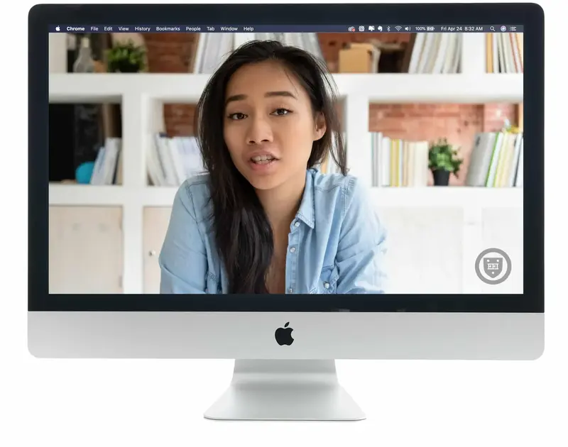 A desktop computer screen showing a young woman in a denim shirt speaking during a video call from a room with bookshelves.