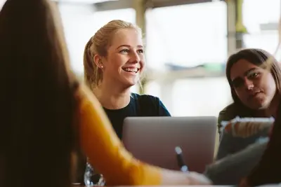 A smiling student sits with classmates around a laptop in a bright classroom