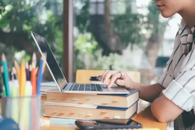 A student typing on a laptop propped on a stack of books, beside a cup of colored pencils by a window