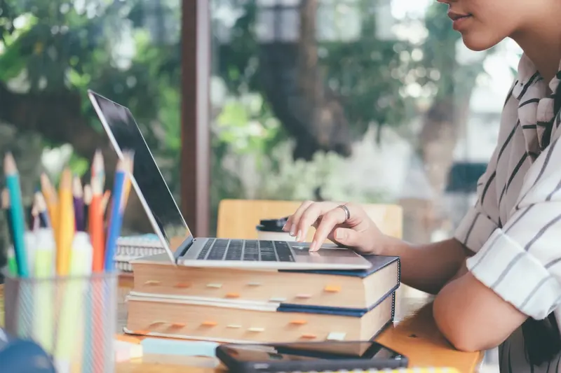 A student typing on a laptop propped on a stack of books, beside a cup of colored pencils by a window