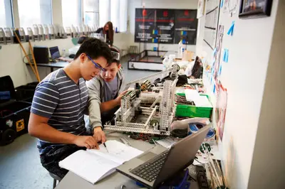 Two students in safety glasses work on notes and a metal robot beside a laptop in a classroom lab.