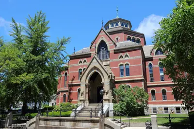 Red brick Gothic-style university building with arched entrance and stone steps under a blue sky, framed by trees