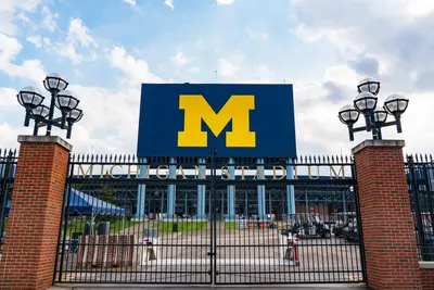 Closed iron gates beneath a sign reading "Michigan Stadium" and a blue scoreboard with a yellow block M.