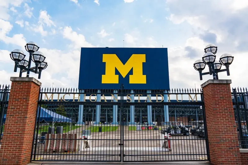 Closed iron gates beneath a sign reading "Michigan Stadium" and a blue scoreboard with a yellow block M.