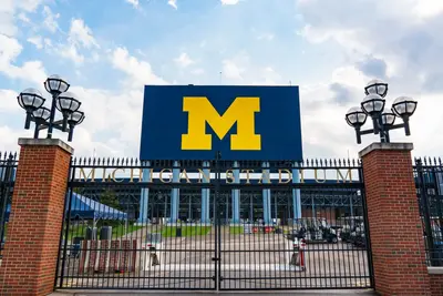 Closed iron gates beneath a sign reading "Michigan Stadium" and a blue scoreboard with a yellow block M.