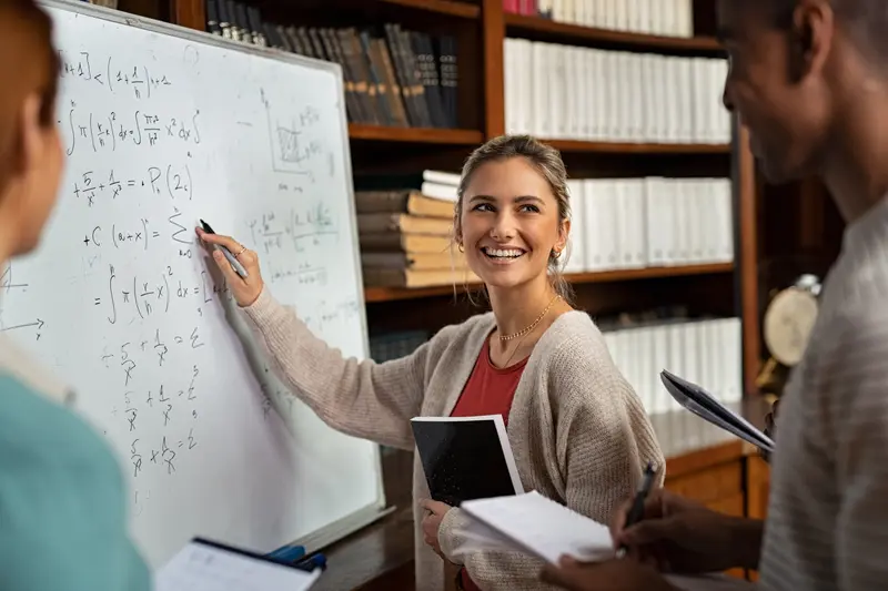 Smiling student writes math equations on a whiteboard while two classmates with notebooks look on in a library.