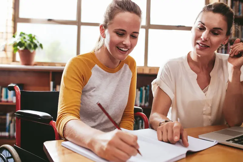 A smiling student in a wheelchair writes in a notebook while a woman beside her looks on in a library.
