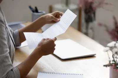 A person seated at a desk holding two printed letters, with a closed laptop and notebook nearby.