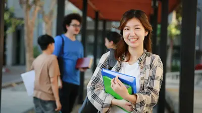 A smiling student holding green and blue folders on a campus walkway, with classmates talking in the background.