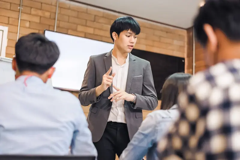 A young man in a grey blazer counts on his fingers while speaking to seated students in a classroom