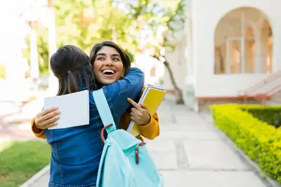 Two students hugging outside on campus, one smiling while holding notebooks and a backpack
