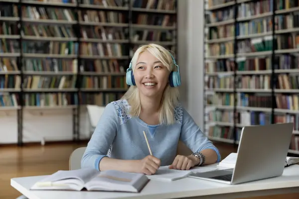 Smiling student wearing blue headphones studies with notebooks and a laptop at a library table lined with bookshelves