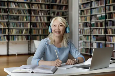 Smiling student wearing blue headphones sits with a pencil, notebook and laptop at a library table lined with bookshelves