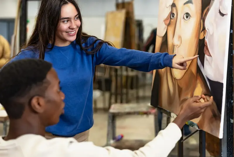 A smiling student points at a large portrait painting on an easel as another student works on it in an art studio.